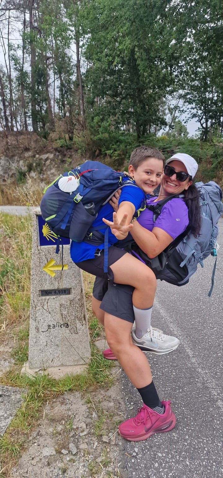 Chiara con il bambino in braccio e gli zaini, accanto a un cippo del Cammino con una freccia gialla.
